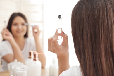 Beautiful woman applying oil onto her eyelashes near mirror indoors