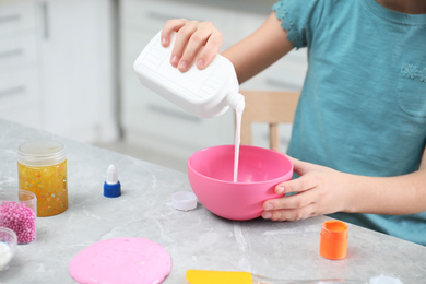 Little girl pouring glue into bowl at table in kitchen, closeup. DIY slime toy