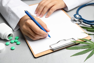 Photo of Female doctor with clipboard, pills and hemp leaf at grey table, closeup