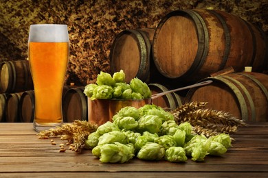 Glass of tasty light beer, fresh hops and wheat spikes on wooden table in cellar