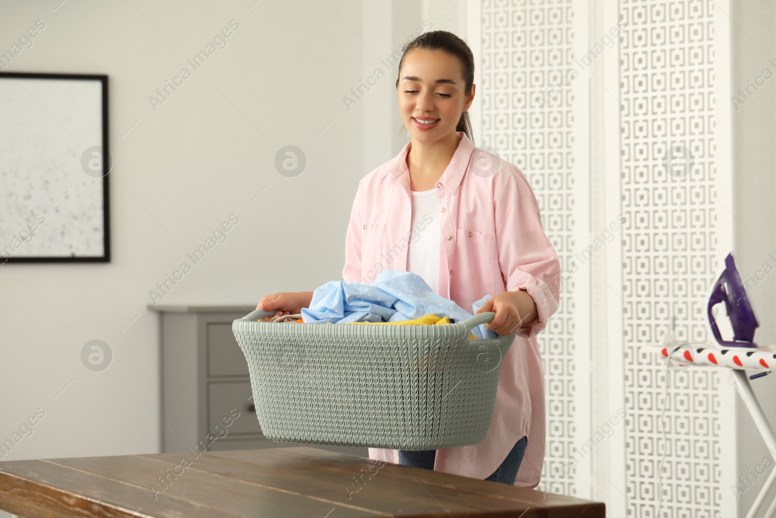 Photo of Young woman putting basket full of clean laundry on wooden table indoors