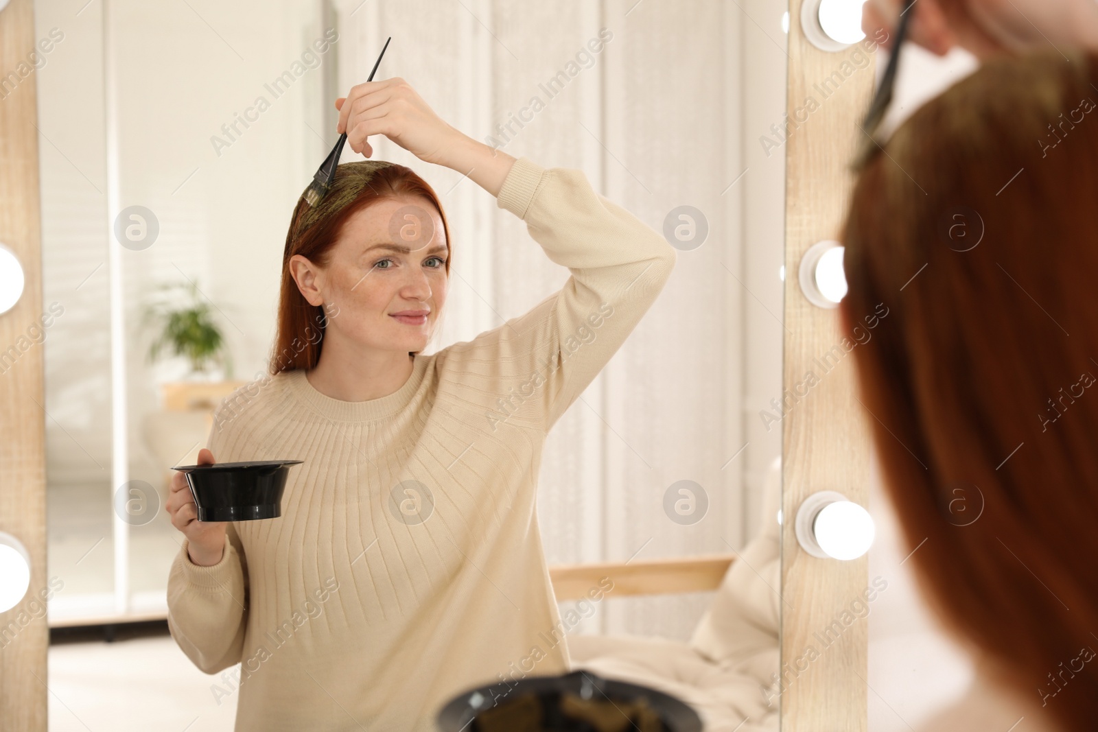Photo of Young woman dyeing her hair with henna near mirror