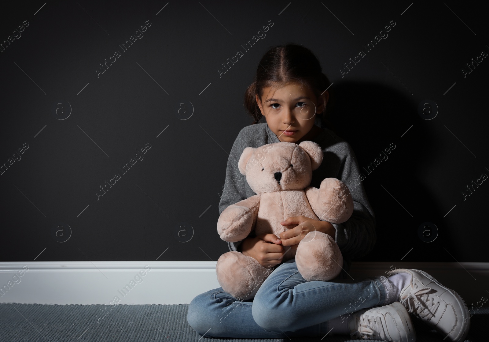 Photo of Sad little girl with teddy bear near black wall, space for text. Domestic violence concept