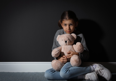 Photo of Sad little girl with teddy bear near black wall, space for text. Domestic violence concept
