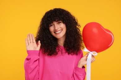Photo of Young woman holding red heart shaped balloon and waving hello on yellow background. Love at distance concept