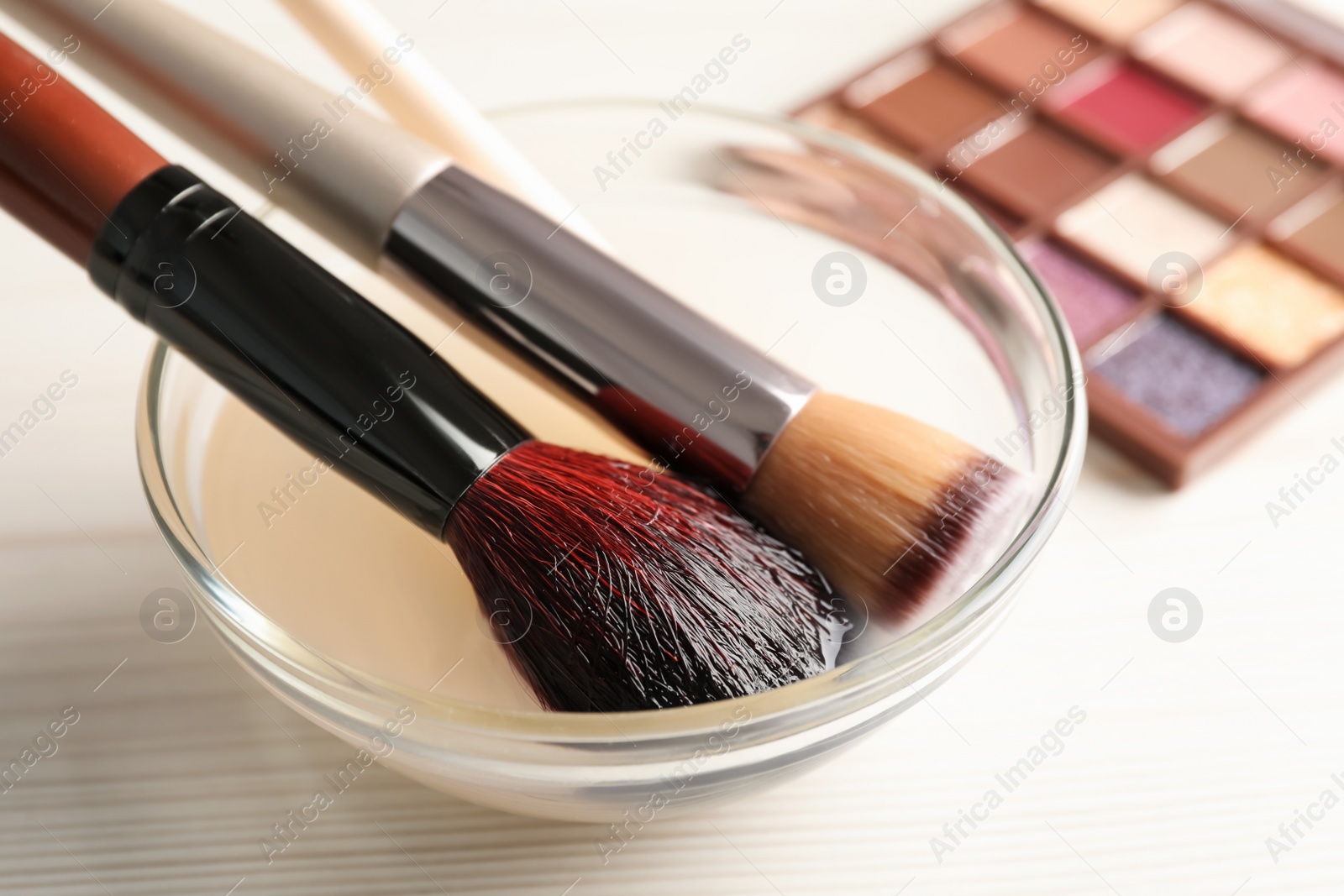 Photo of Cleaning makeup brushes in bowl with special liquid on white wooden table, closeup