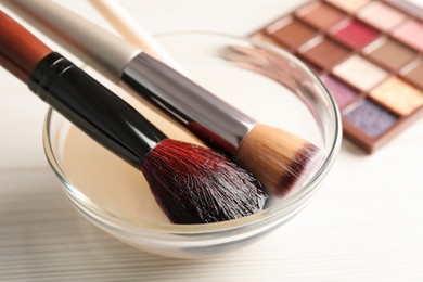 Photo of Cleaning makeup brushes in bowl with special liquid on white wooden table, closeup