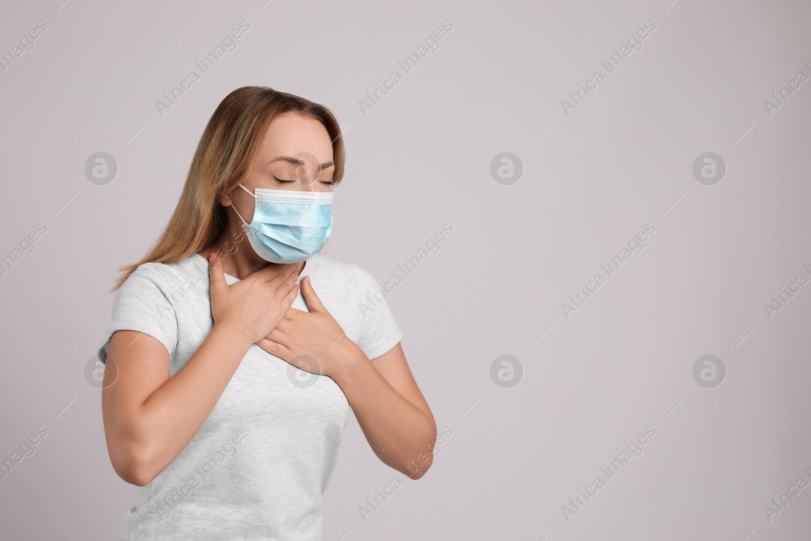 Photo of Young woman in medical mask suffering from pain during breathing on light grey background, space for text