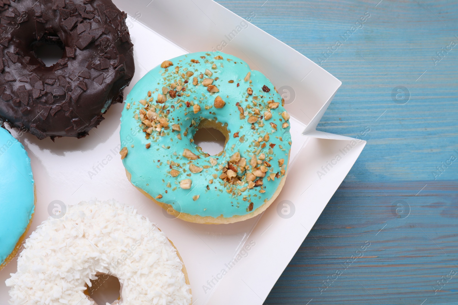 Photo of Box with different tasty glazed donuts on light blue wooden table, closeup