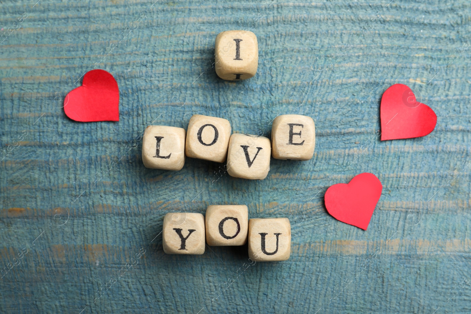 Photo of Phrase I Love You made of decorative cubes and paper hearts on light blue wooden background, flat lay