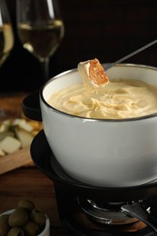 Dipping piece of bread into fondue pot with melted cheese at table, closeup