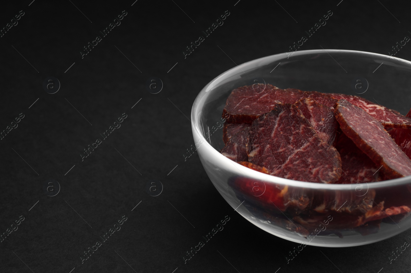 Photo of Bowl with pieces of delicious beef jerky on black table, closeup. Space for text