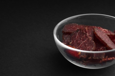 Photo of Bowl with pieces of delicious beef jerky on black table, closeup. Space for text