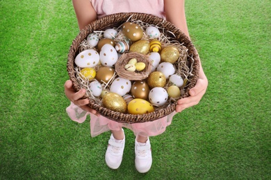 Photo of Little girl holding wicker basket full of Easter eggs outdoors, closeup
