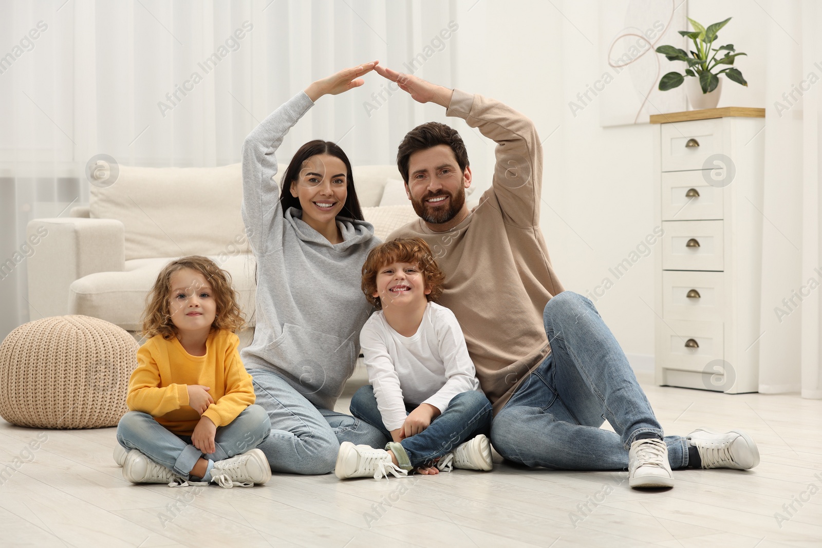 Photo of Family housing concept. Happy woman and her husband forming roof with their hands while sitting with kids on floor at home