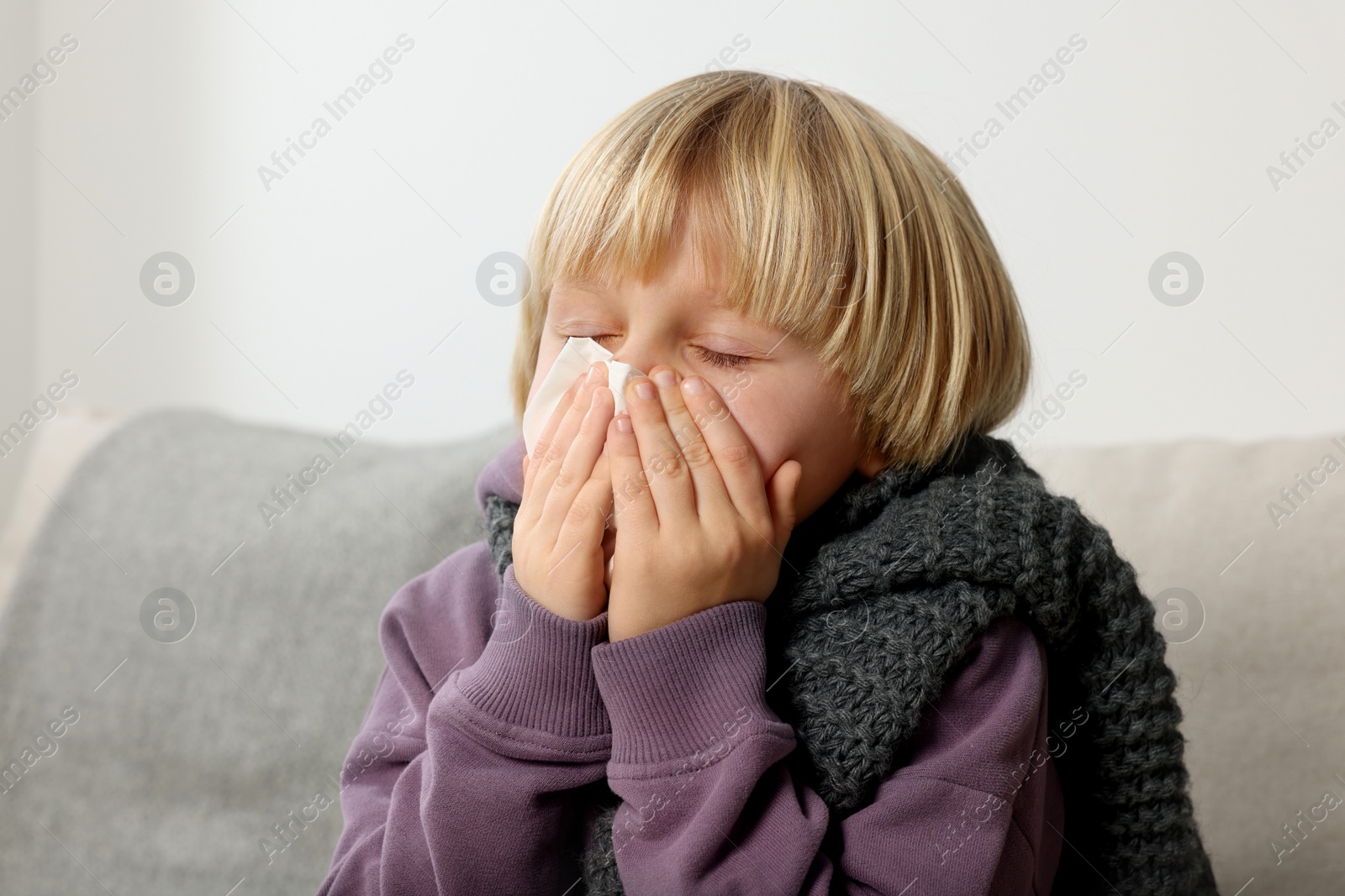 Photo of Boy blowing nose in tissue on sofa in room. Cold symptoms