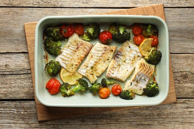 Photo of Pieces of delicious baked cod with vegetables, lemon and spices in dish on wooden table, top view