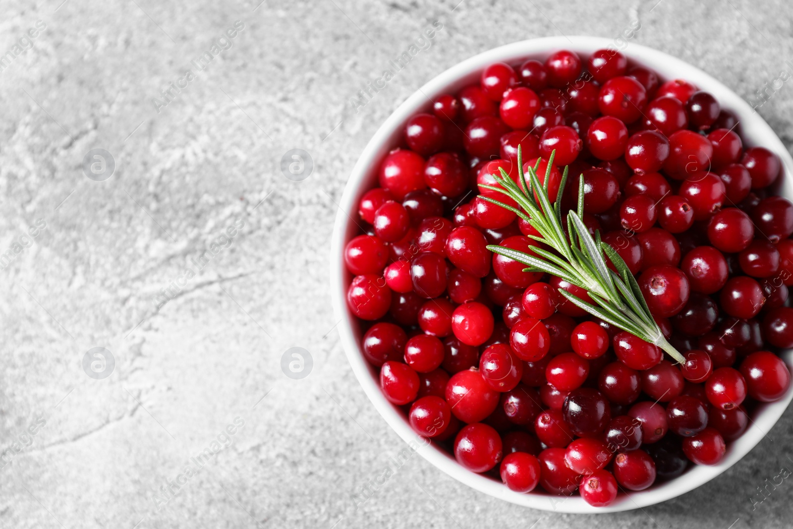 Photo of Fresh ripe cranberries and rosemary in bowl on grey table, top view. Space for text