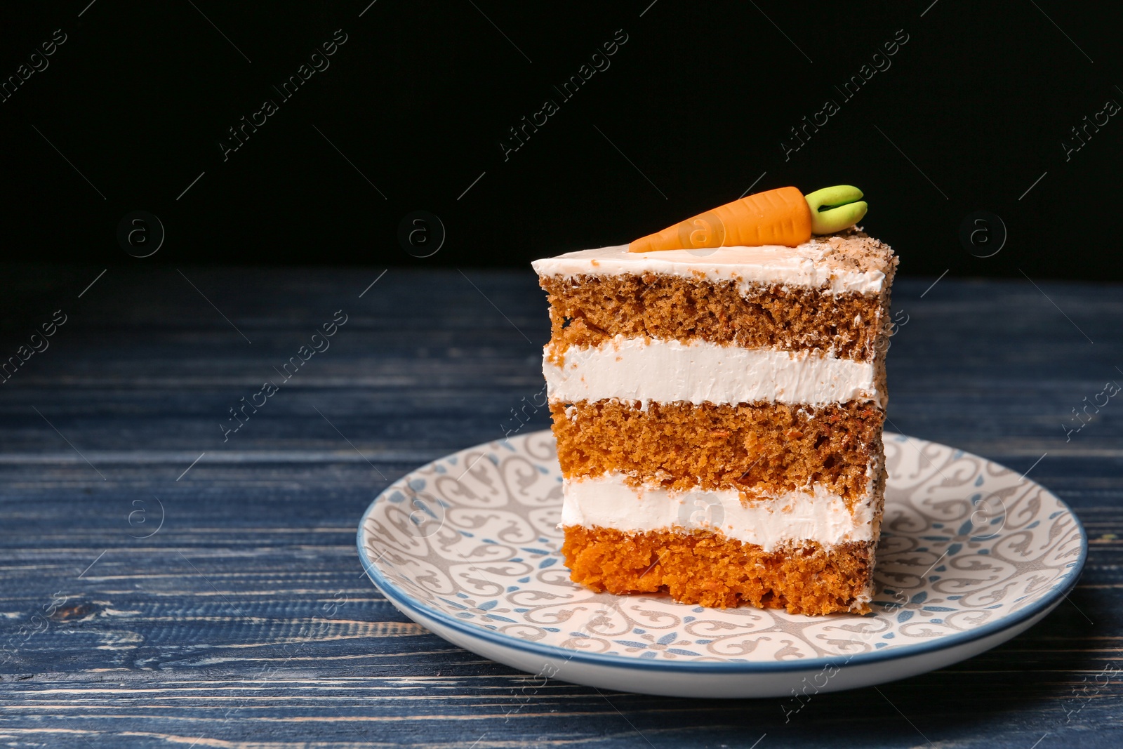 Photo of Piece of sweet carrot cake with delicious cream on blue wooden table against dark background. Space for text