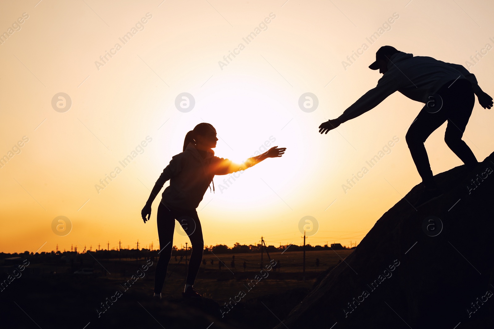 Photo of Silhouettes of man and woman helping each other to climb on hill against sunset