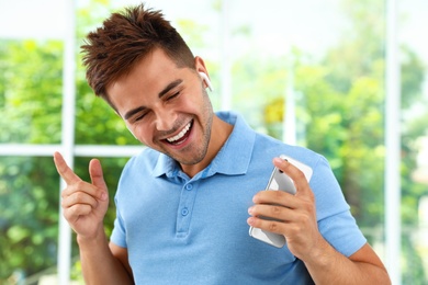 Photo of Happy young man with smartphone listening to music through wireless earphones indoors