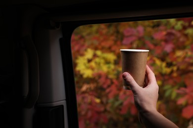 Photo of Woman holding takeaway cardboard coffee cup in car among autumn forest, closeup