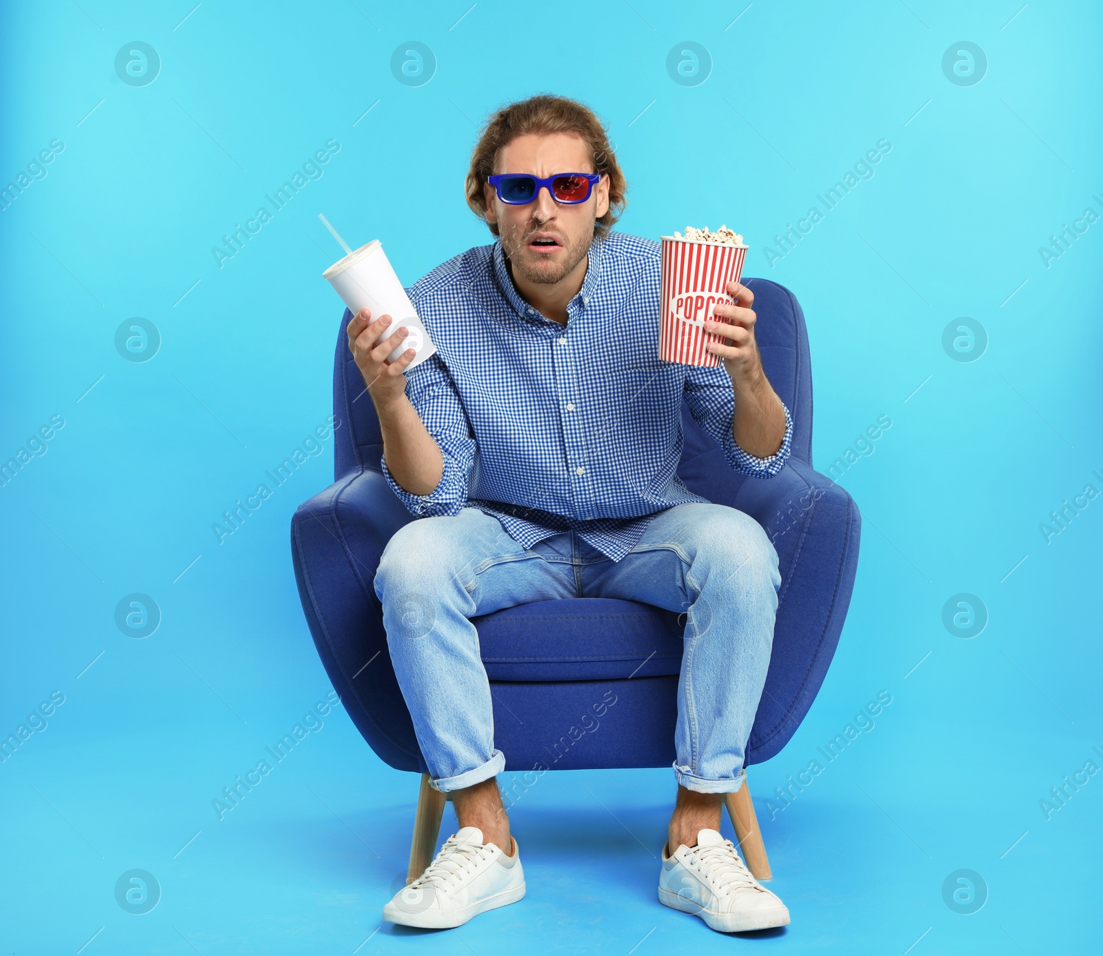 Photo of Emotional man with 3D glasses, popcorn and beverage sitting in armchair during cinema show on color background