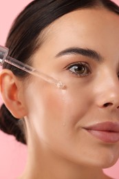Photo of Beautiful young woman applying serum onto her face on pink background, closeup
