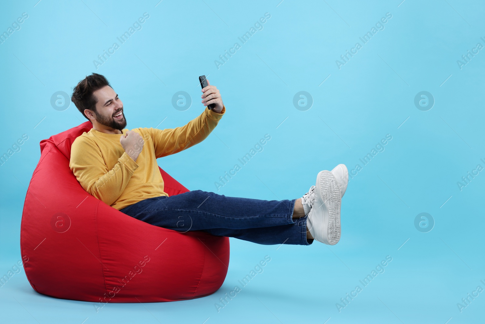 Photo of Happy young man using smartphone on bean bag chair against light blue background. Space for text