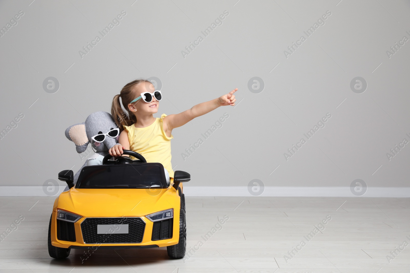 Photo of Cute little girl with toy elephant driving children's car near grey wall indoors. Space for text