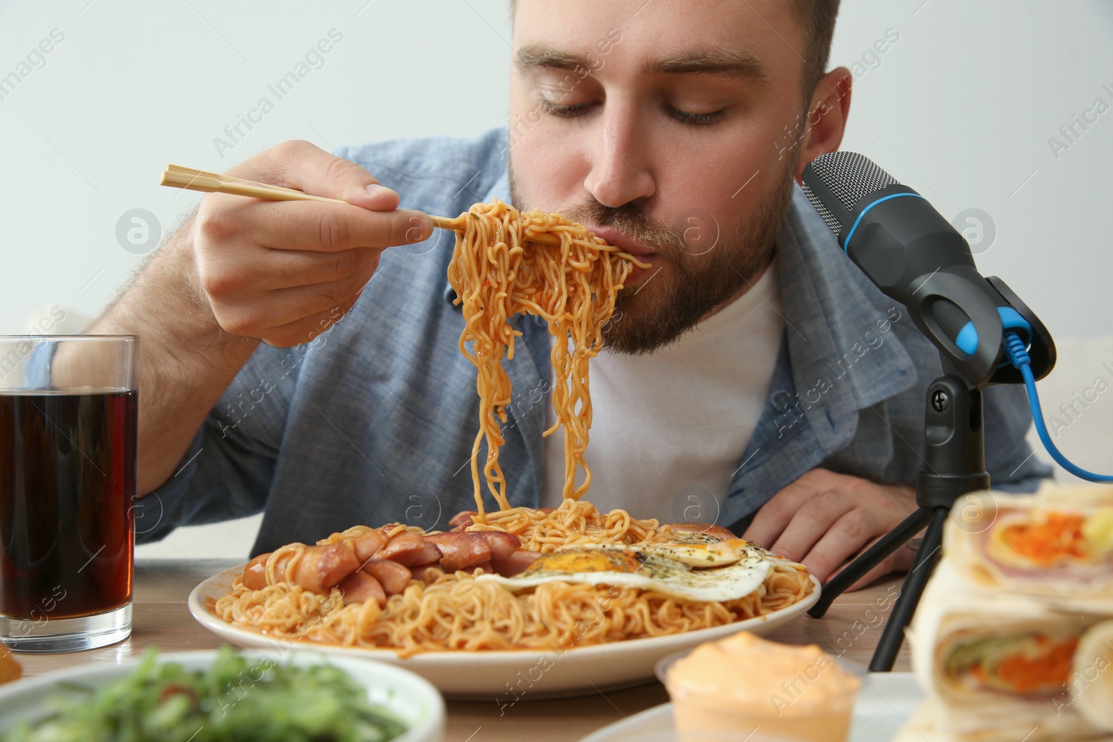 Photo of Food blogger eating in front of microphone at table against light background. Mukbang vlog