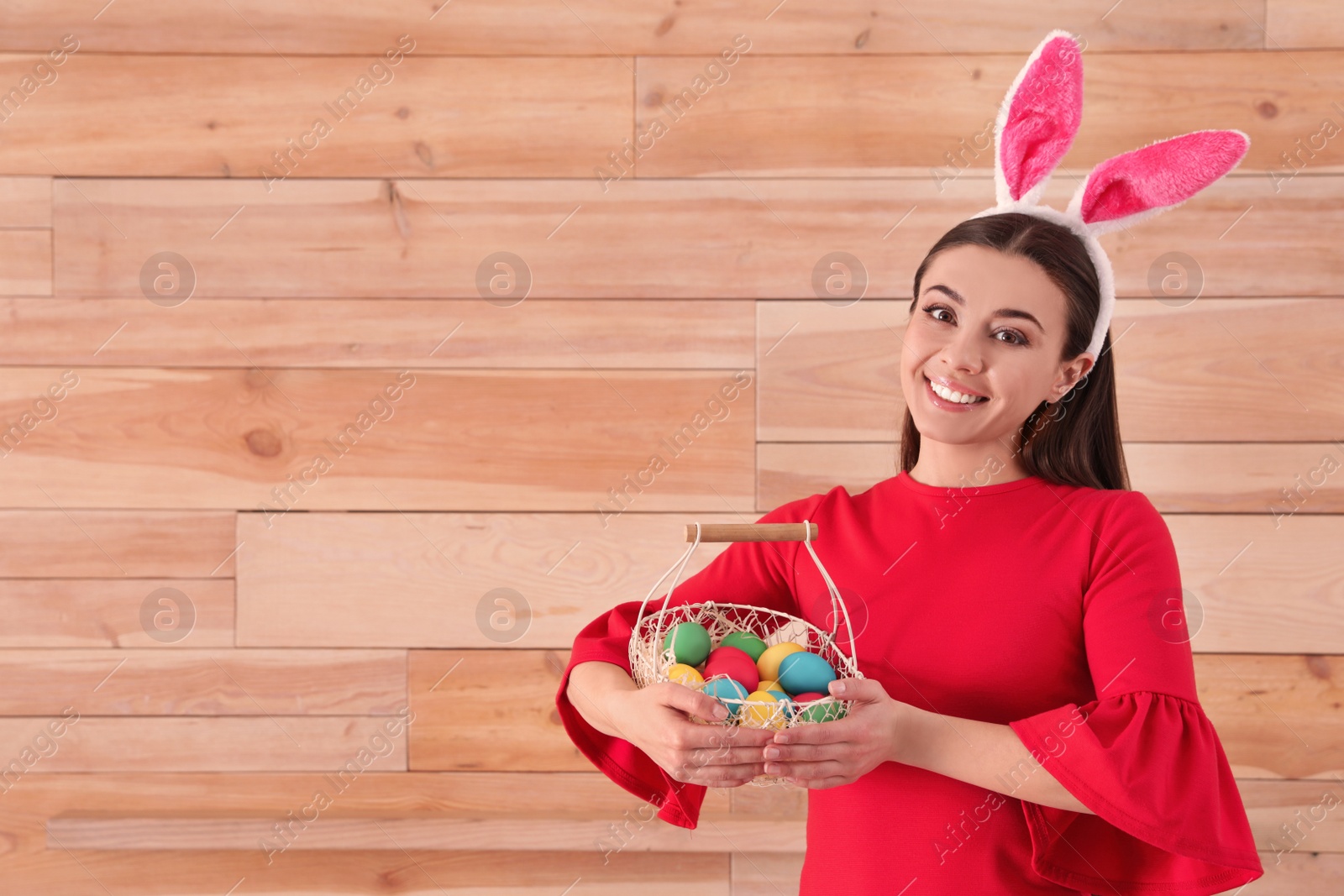 Photo of Beautiful woman in bunny ears headband holding basket with Easter eggs on wooden background, space for text