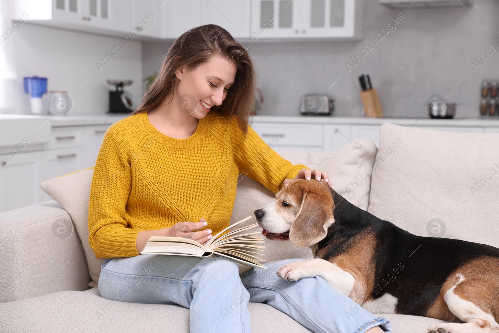 Photo of Happy young woman reading book near her cute Beagle dog on couch at home. Lovely pet