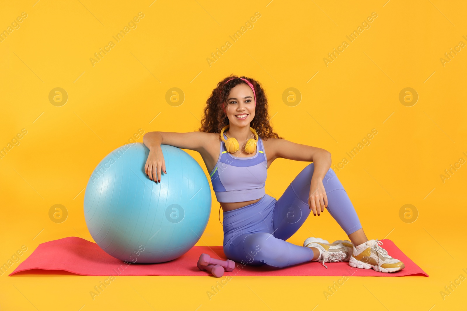 Photo of Beautiful African American woman with headphones and sports equipments sitting on yoga mat against yellow background