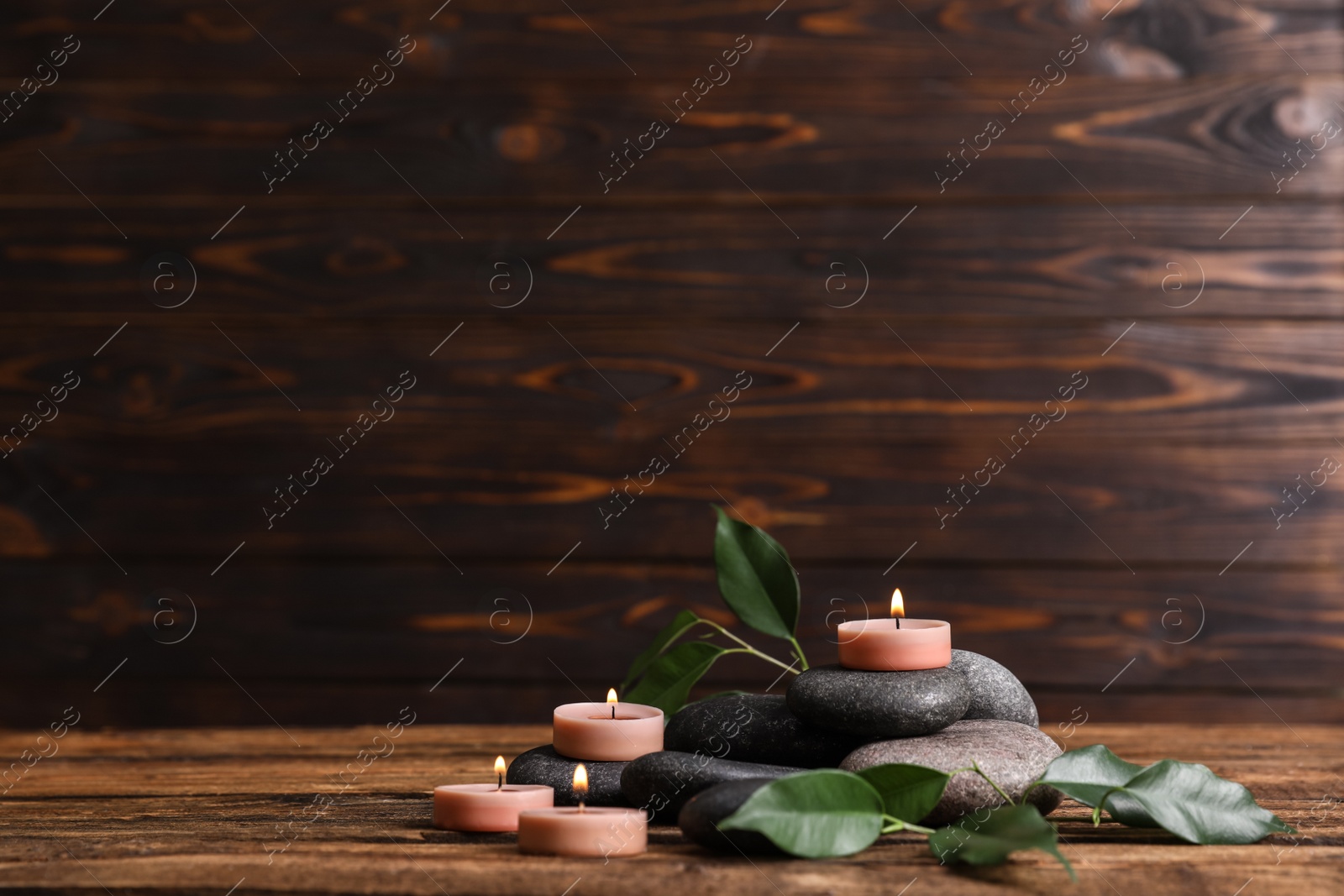 Photo of Composition of spa stones, green leaves and burning candles on wooden table, space for text