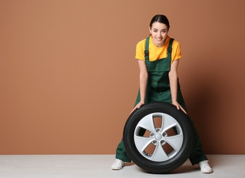 Female mechanic in uniform with car tire on color wall background