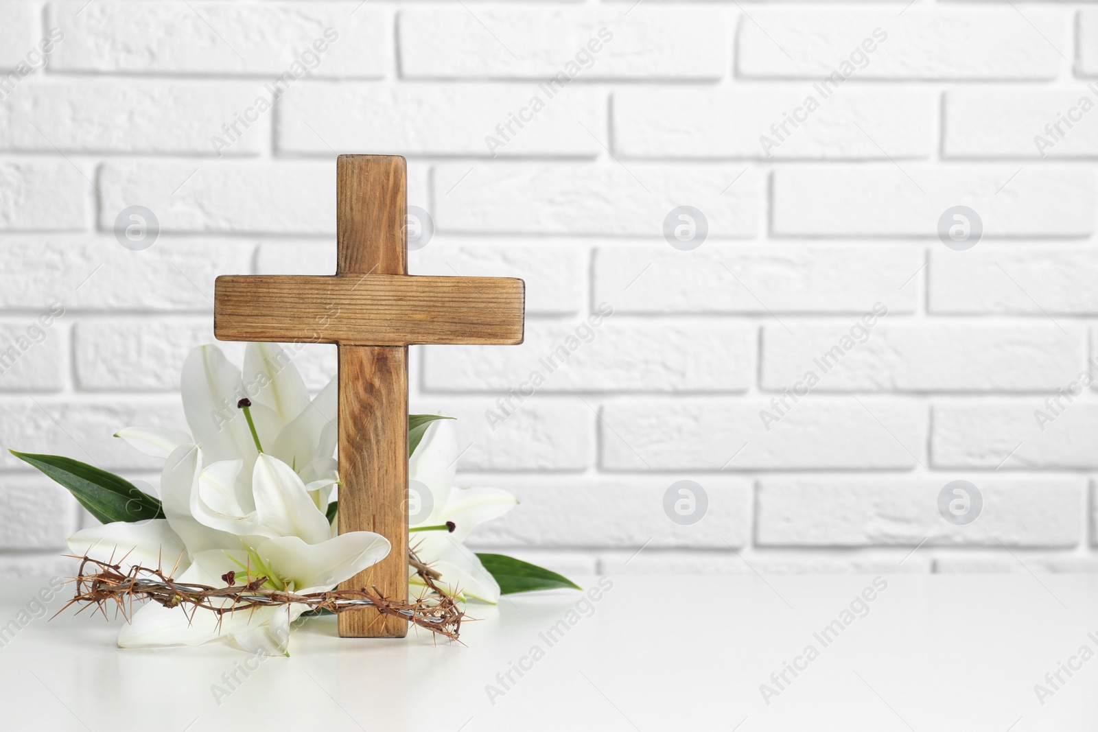 Photo of Wooden cross, crown of thorns and blossom lilies on table against brick wall, space for text