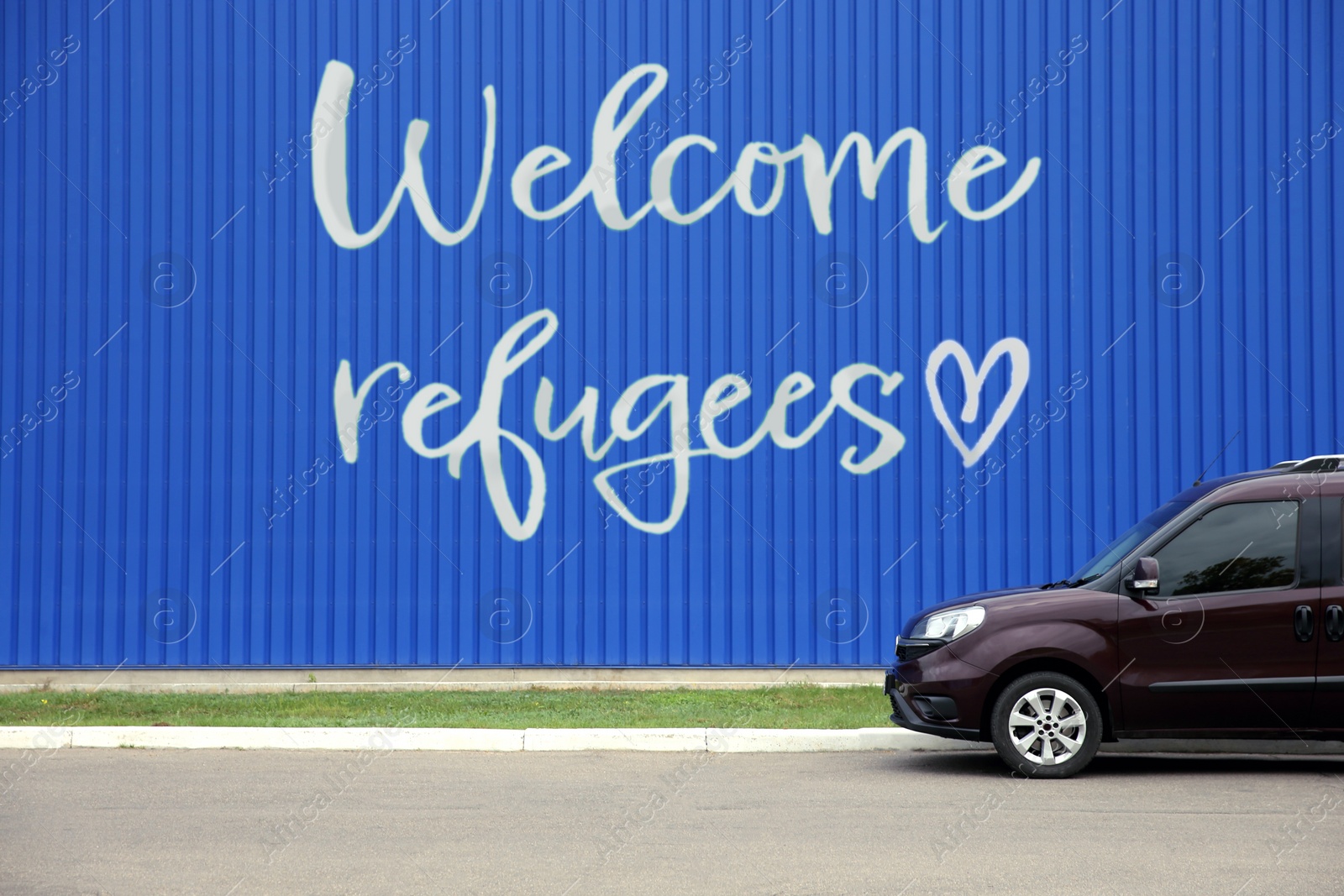 Image of Car parked near blue wall with phrase WELCOME REFUGEES outdoors