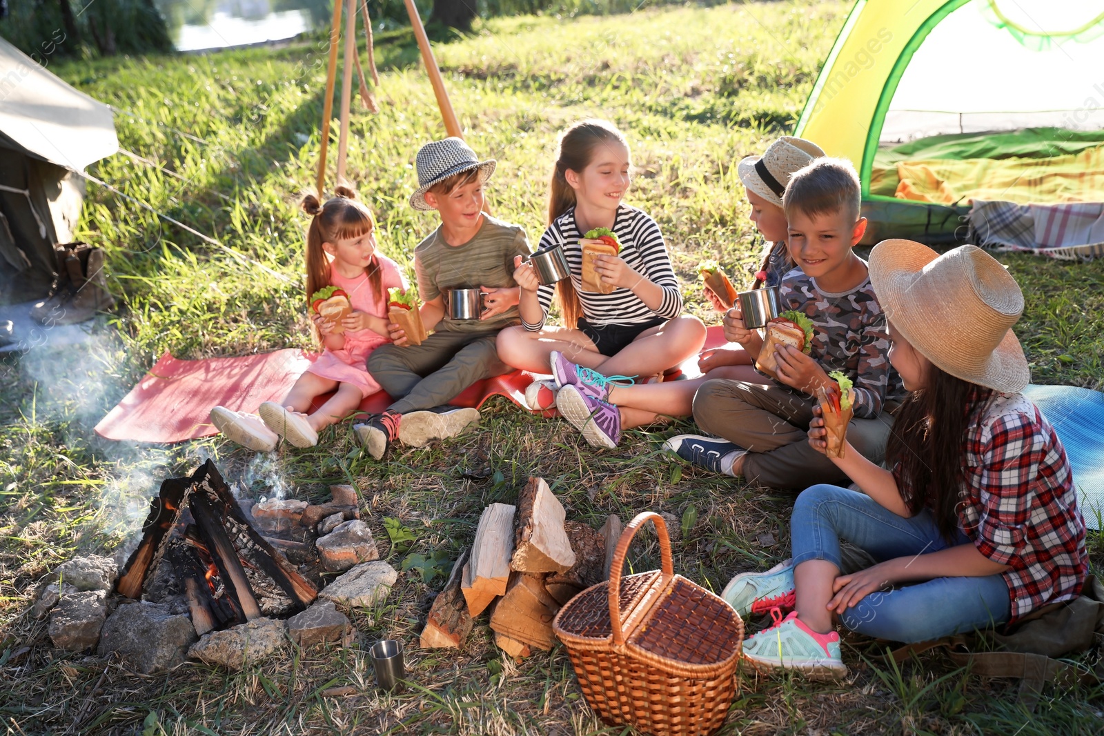 Photo of Little children eating sandwiches near bonfire and tent. Summer camp