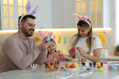 Photo of Easter celebration. Happy family with bunny ears painting eggs at white marble table in kitchen
