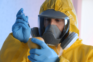 Scientist in chemical protective suit injecting apple on color background, closeup