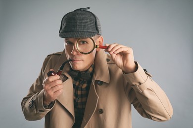 Male detective with magnifying glass smoking pipe on grey background