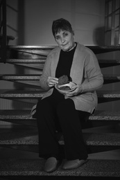 Poor senior woman with bread sitting on stairs indoors, black and white effect