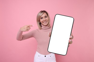 Happy woman pointing at mobile phone with blank screen on pink background. Mockup for design