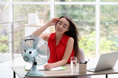 Photo of Young woman enjoying air flow from fan at workplace. Summer heat