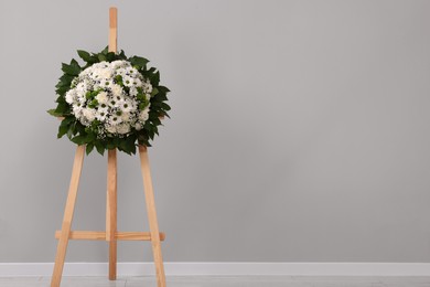Photo of Funeral wreath of flowers on wooden stand near grey wall indoors. Space for text