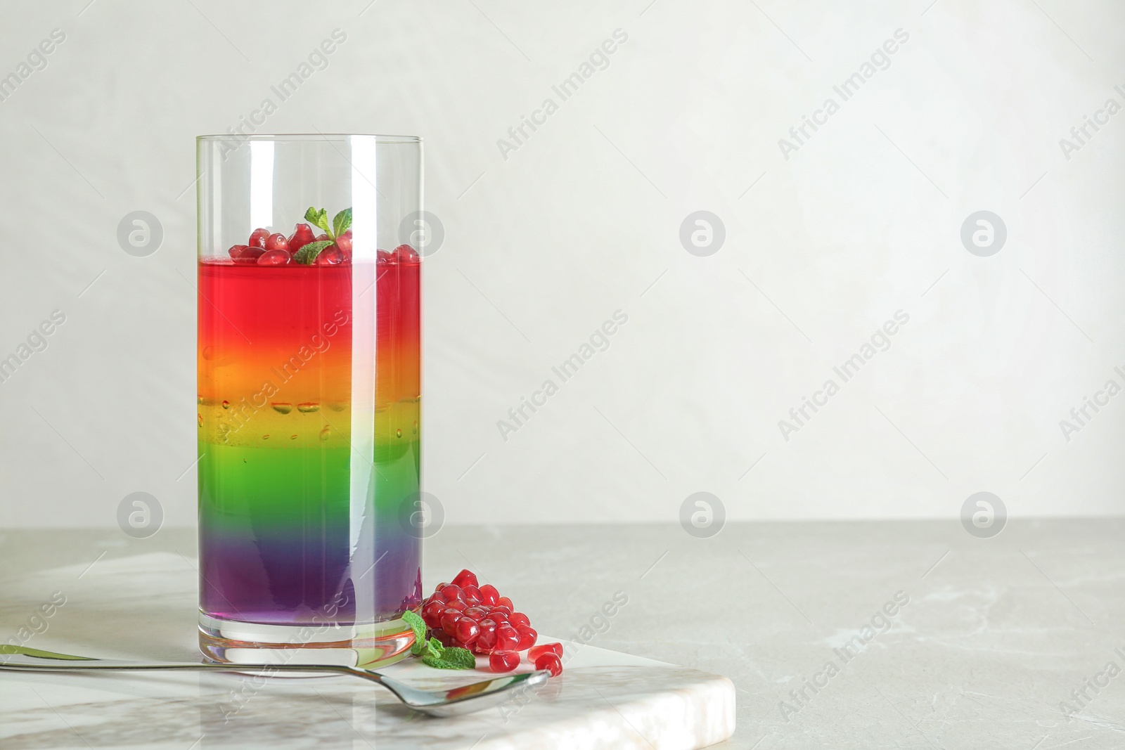 Photo of Board with glass of colorful jelly, spoon and pomegranate seeds on light table. Space for text