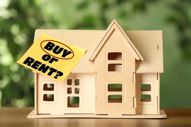 Model of house with sticky note Buy Or Rent on wooden table against blurred green background