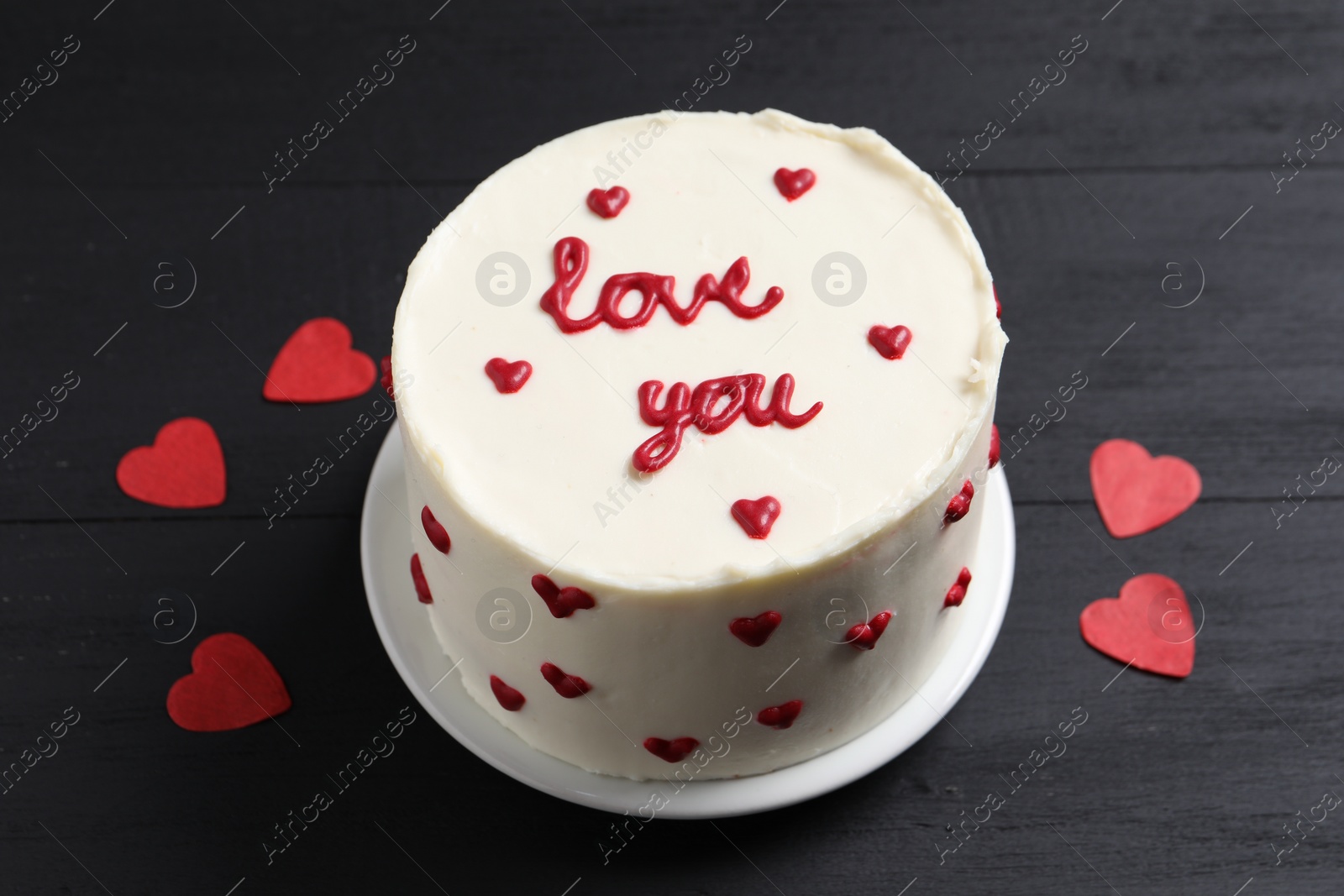 Photo of Bento cake with text Love You and paper hearts on black wooden table. St. Valentine's day surprise