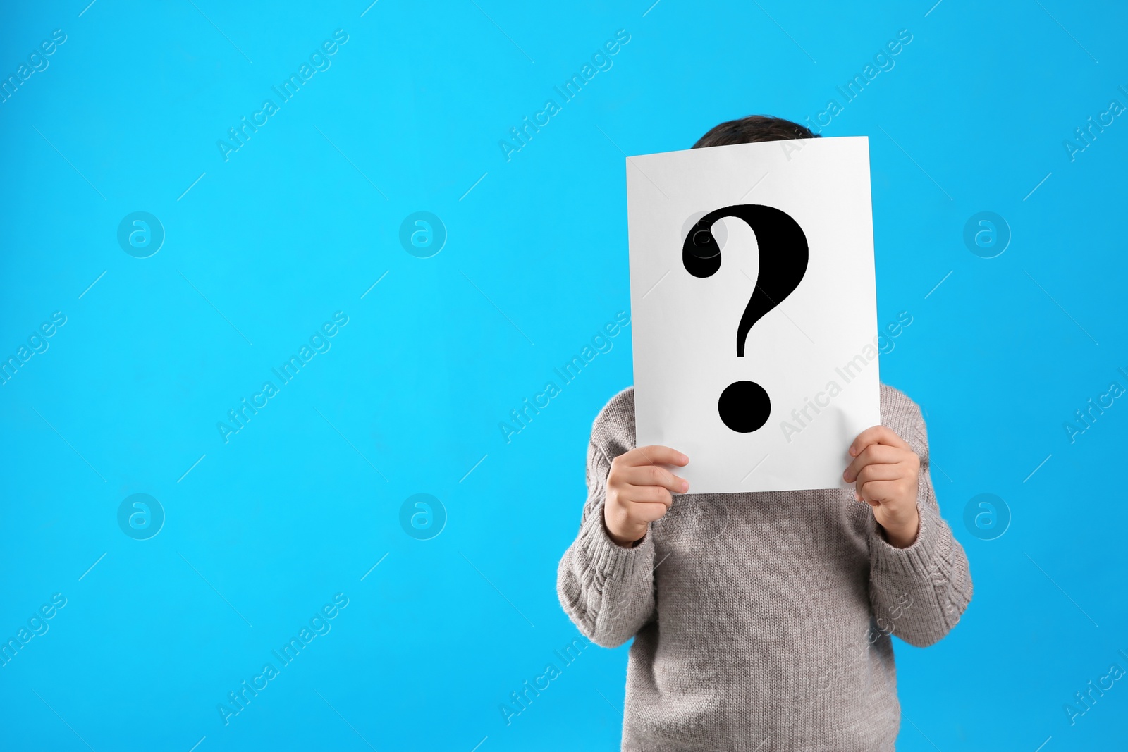 Photo of Little boy holding paper with question mark on light blue background, space for text